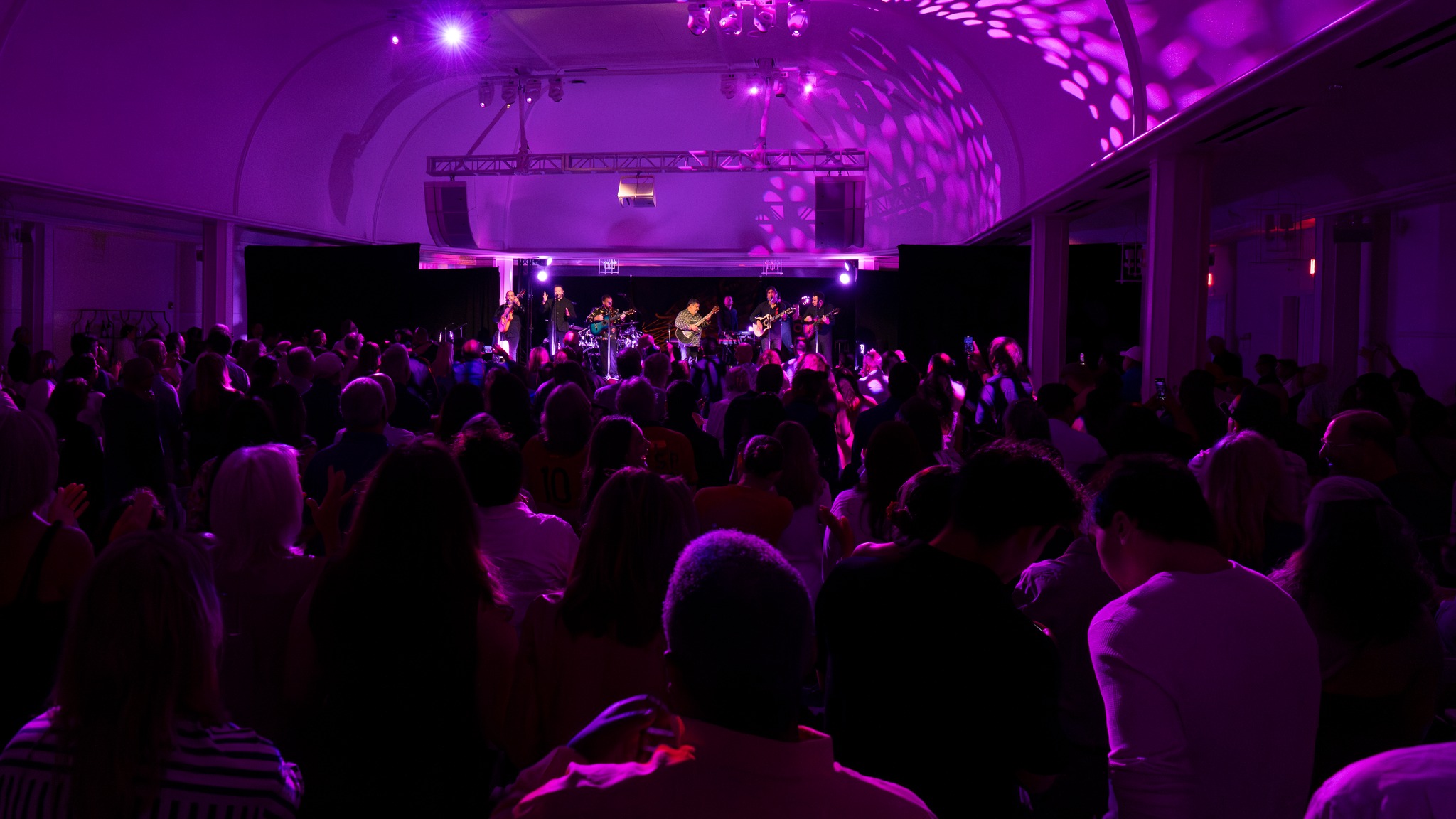 A large crowd watches a live band perform on stage under purple lighting in a spacious indoor venue.